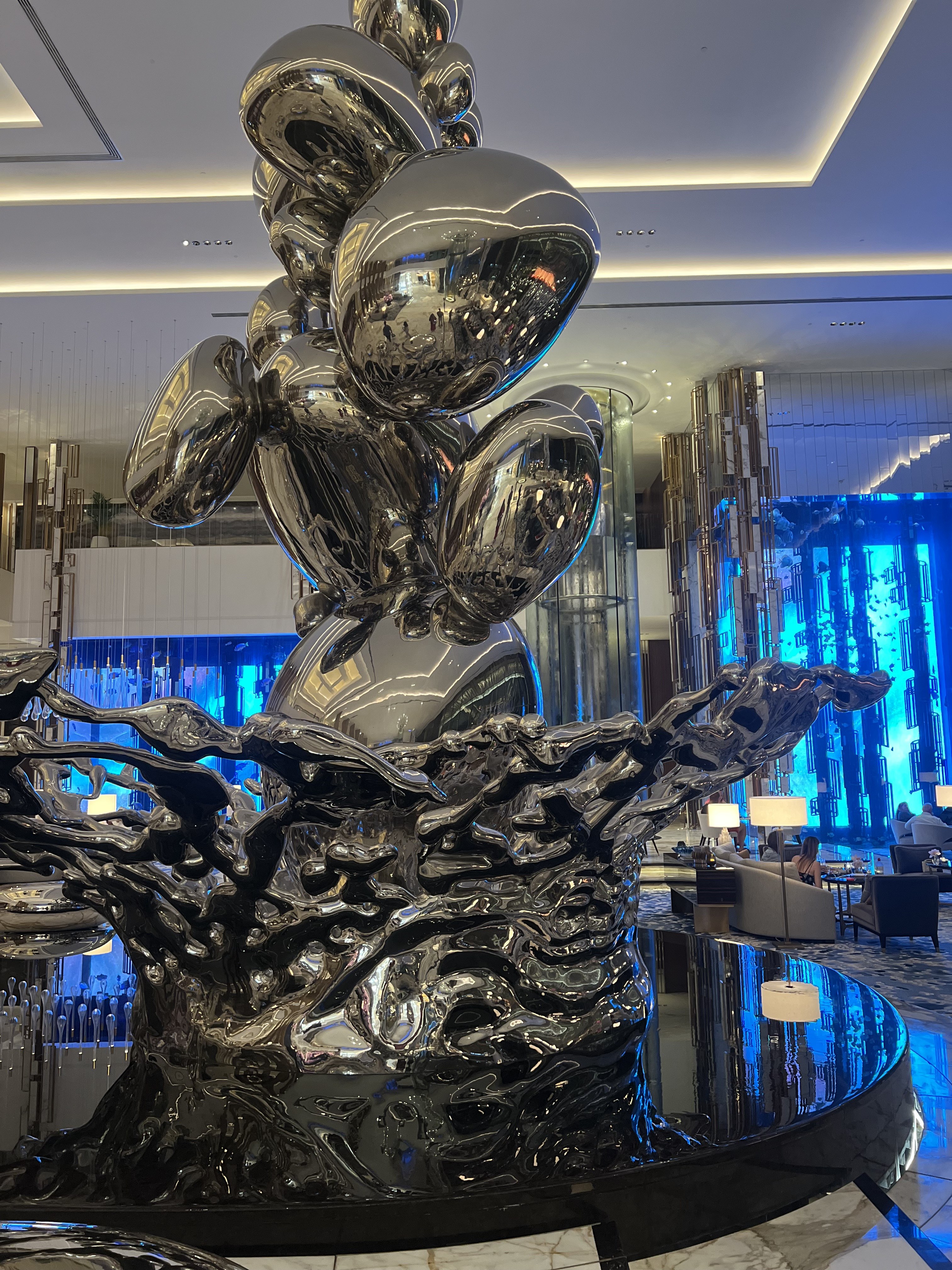 Large polished chrome sculpture rising from black plinth with aquarium wall of deep blue light behind, Atlantis The Royal, Dubai