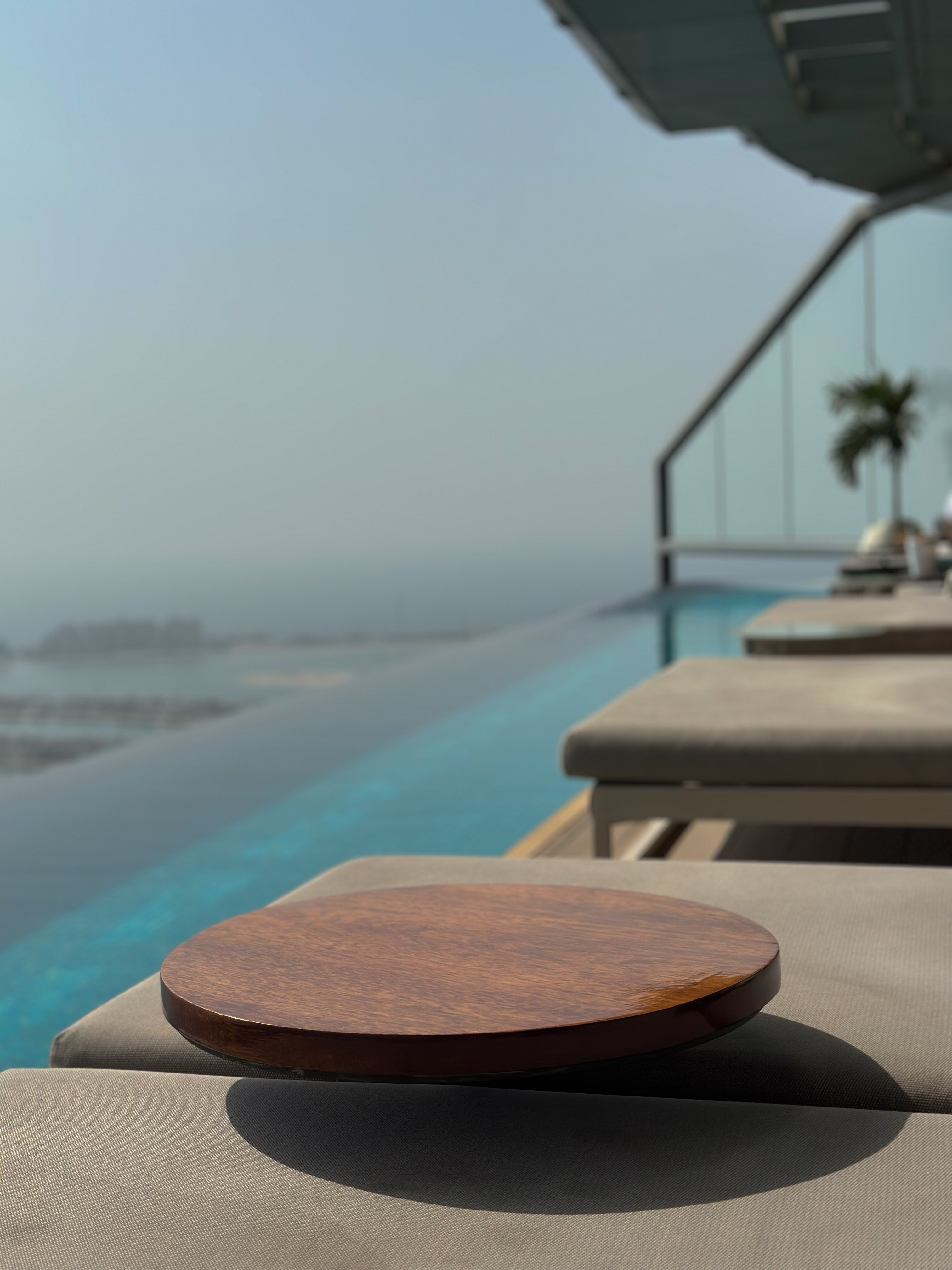 Wooden disc on poolside lounger, infinity pool edge and hazy horizon, AURA, Dubai