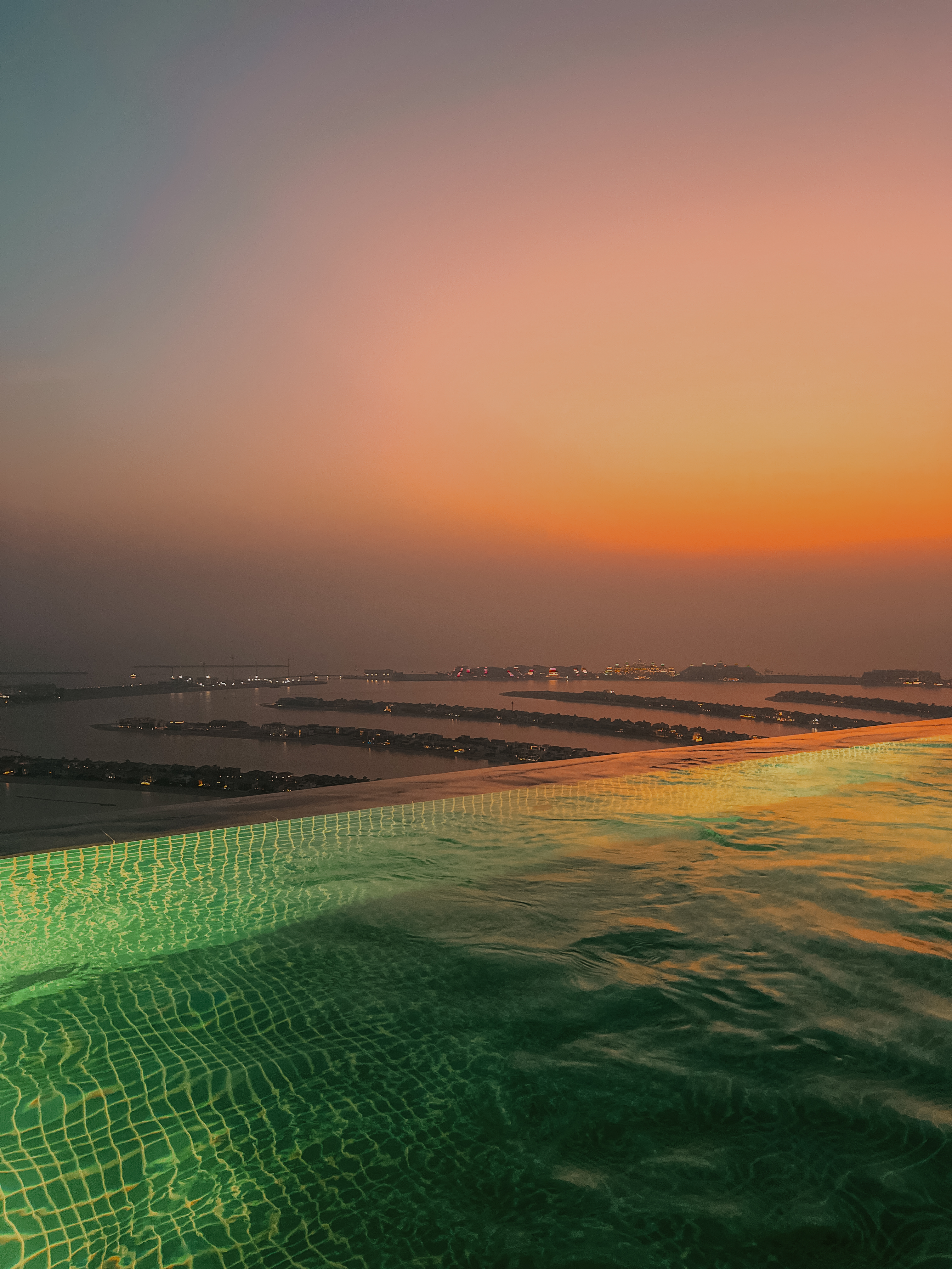 Sunset infinity pool above Palm Jumeirah, pool illuminated green against orange sky, AURA, Dubai