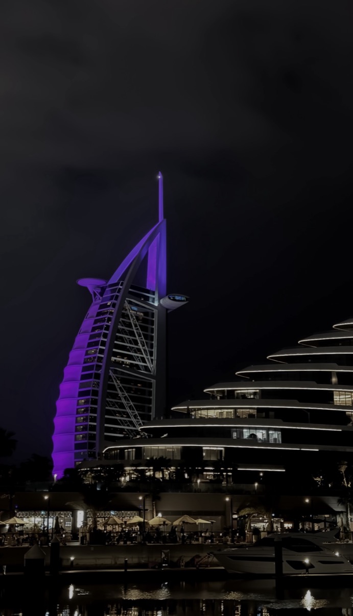 Burj Al Arab lit purple at night with terraced building and water in foreground, Dubai