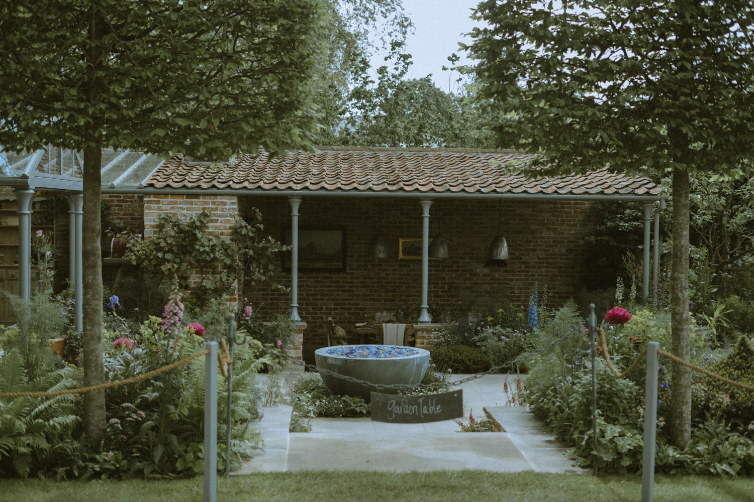 The Savills Garden seen straight on between pleached trees, with the water bowl and Garden Table sign, RHS Chelsea Flower Show, London