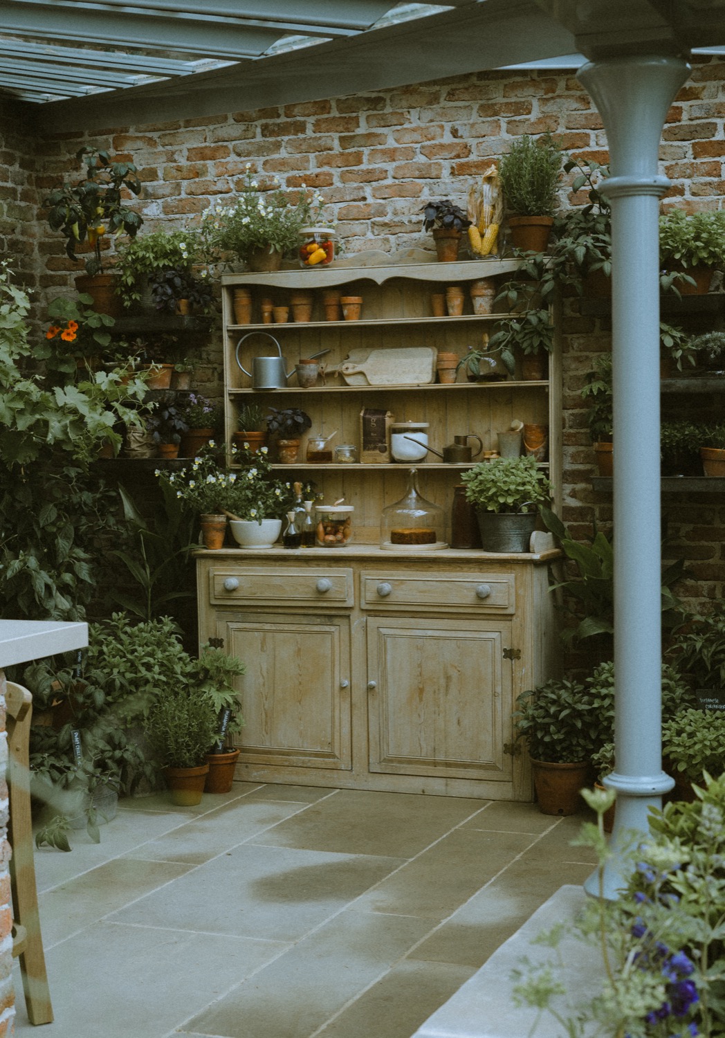 The pine dresser standing against reclaimed brick beside a cast-iron column, The Savills Garden, RHS Chelsea Flower Show, London