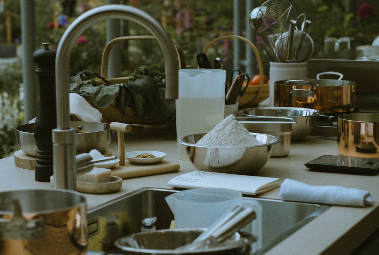 The worktop mid-preparation, with flour in a steel bowl, copper pans and an open notebook, The Savills Garden, RHS Chelsea Flower Show, London