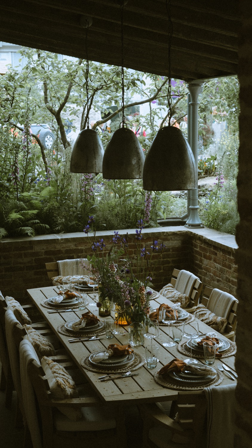 The table laid beneath three concrete pendant lights, with the borders beyond, The Savills Garden, RHS Chelsea Flower Show, London