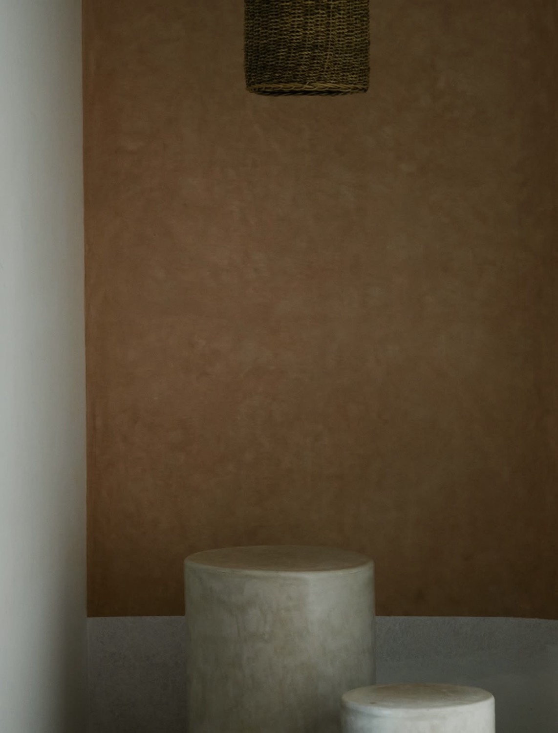 Terracotta plaster wall with woven pendant light shade, concrete cylinder objects on a surface below