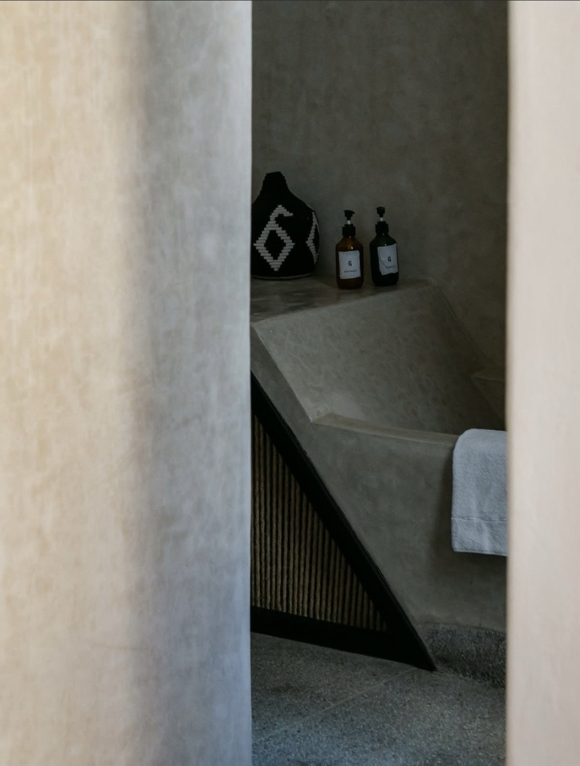 Concrete bathroom alcove glimpsed through a doorway, amber pump bottles and woven basket on shelf