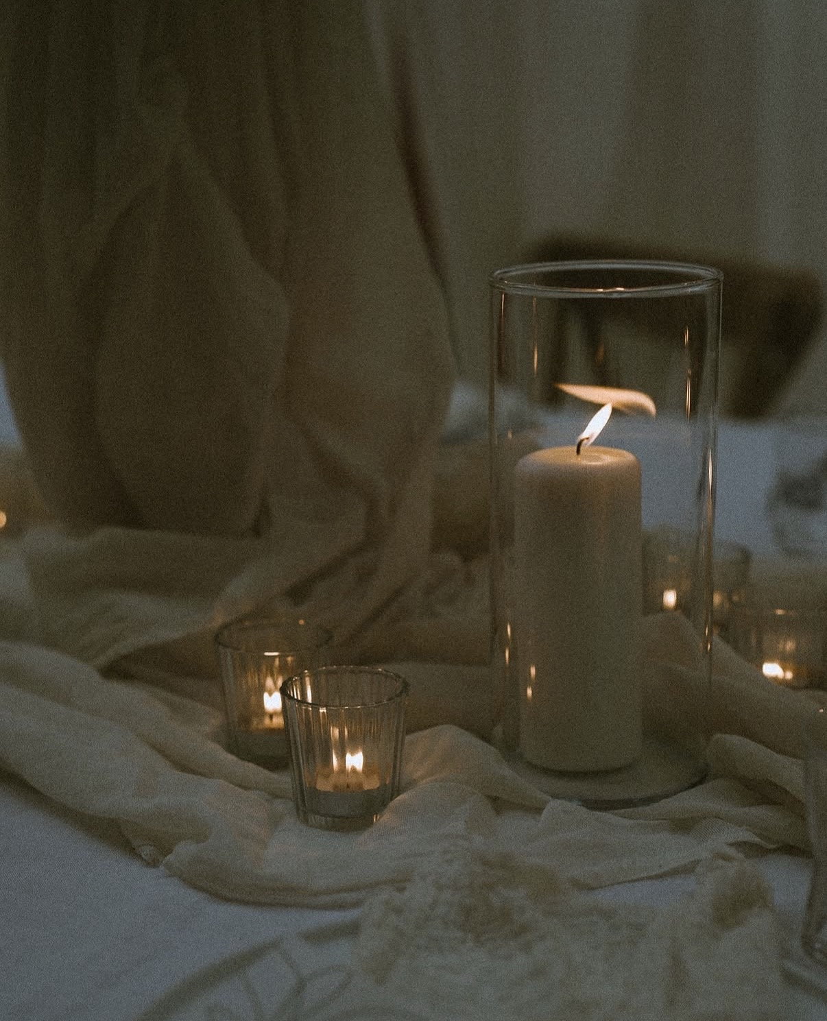 Pillar candle in a glass hurricane vase on white linen, smaller votives beside it
