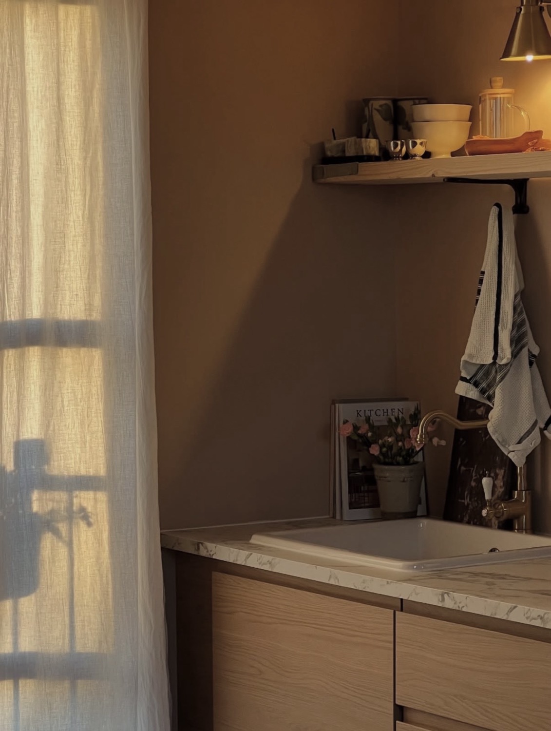 Warm kitchen corner at dusk, terracotta wall, linen curtain with window shadows, brass tap and KITCHEN book on counter