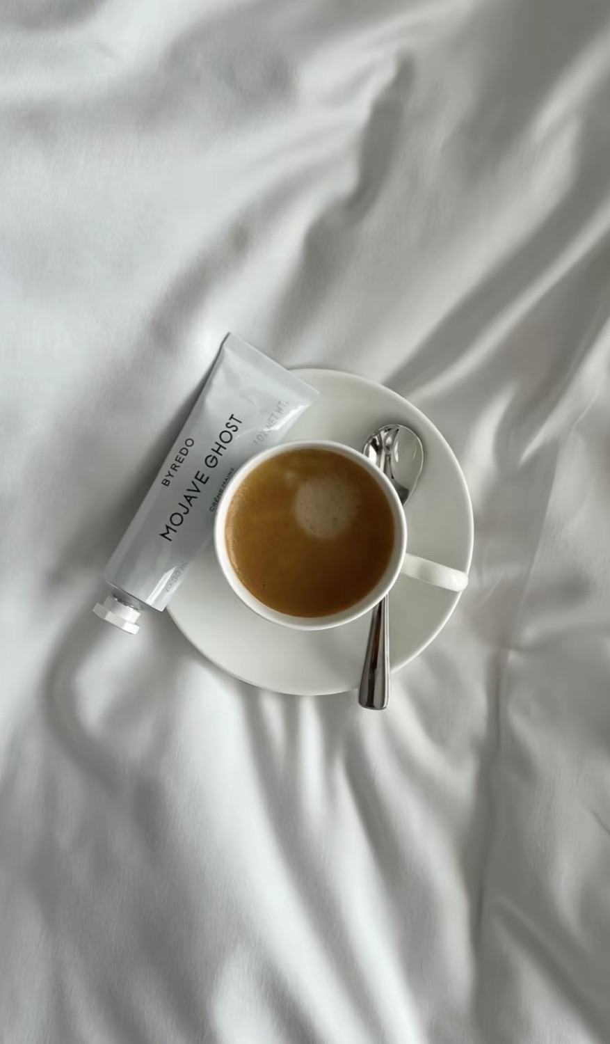 Byredo Mojave Ghost tube beside an espresso cup and saucer on white hotel linen, top-down view