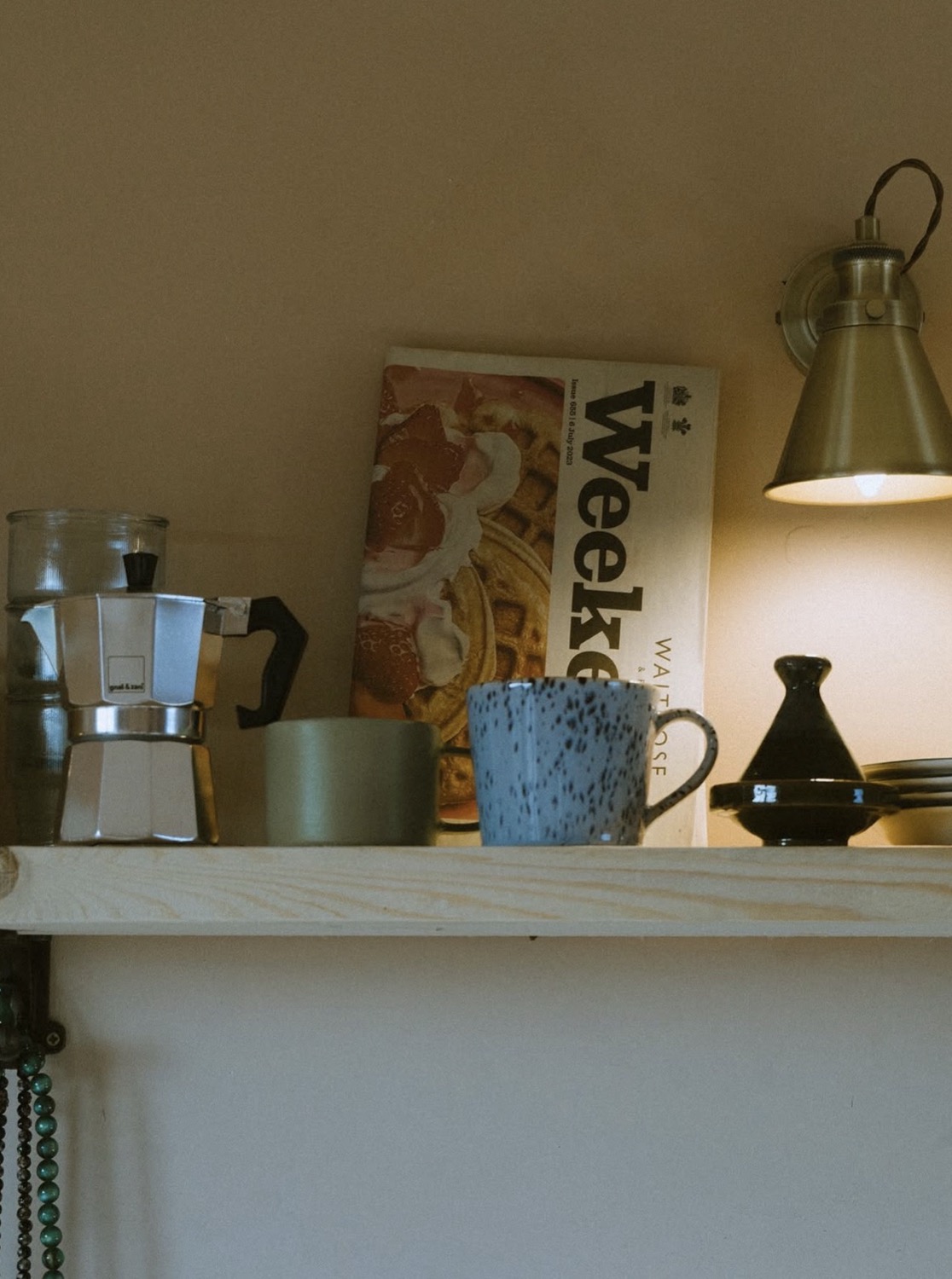 Kitchen shelf with Bialetti moka pot, speckled blue mug, Waitrose Weekend magazine and brass wall lamp