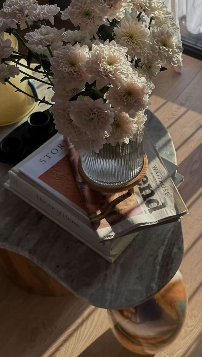 White chrysanthemums in a ribbed glass vase on a marble side table, stacked books and Waitrose Weekend below in sunlight