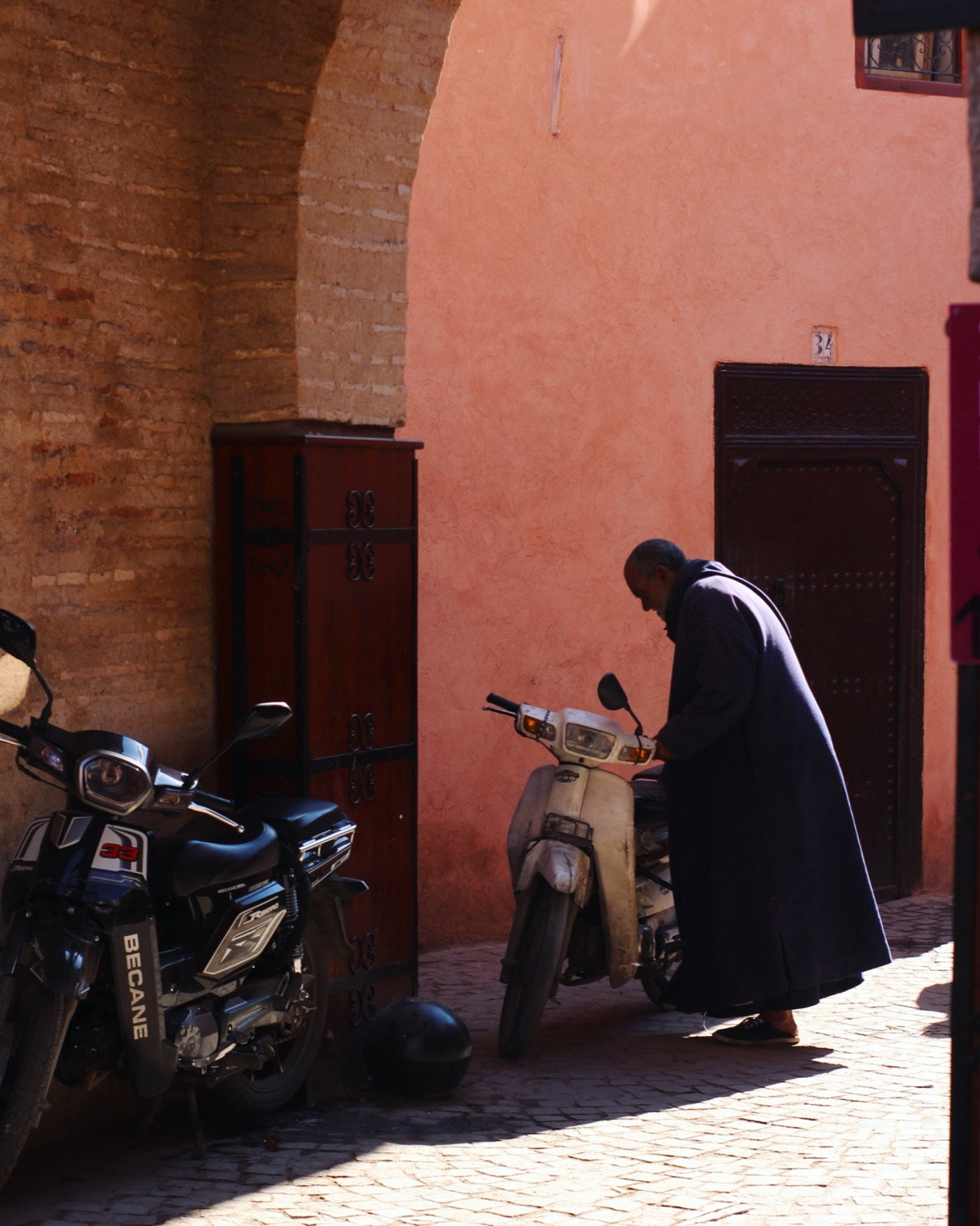 Man in djellaba in a Marrakesh medina alley, scooters and a pink arched wall behind