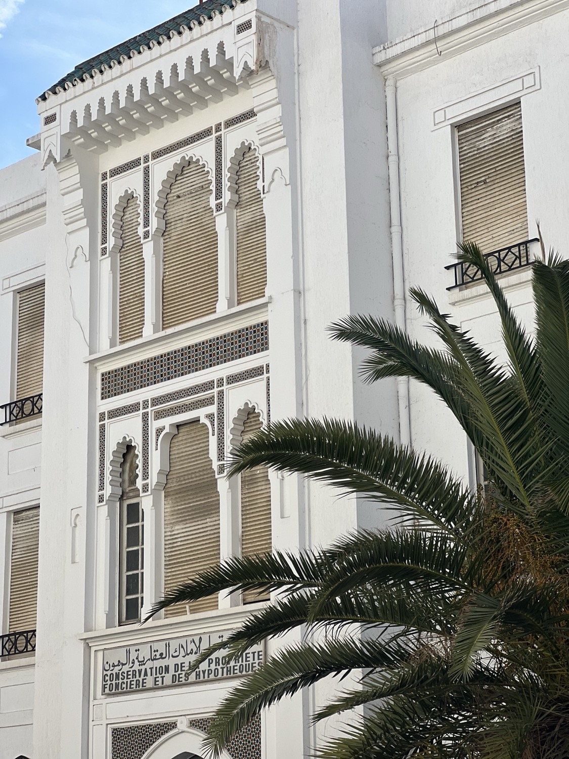 White Moroccan building with ornate carved arch windows and a palm frond in the foreground, Tangier