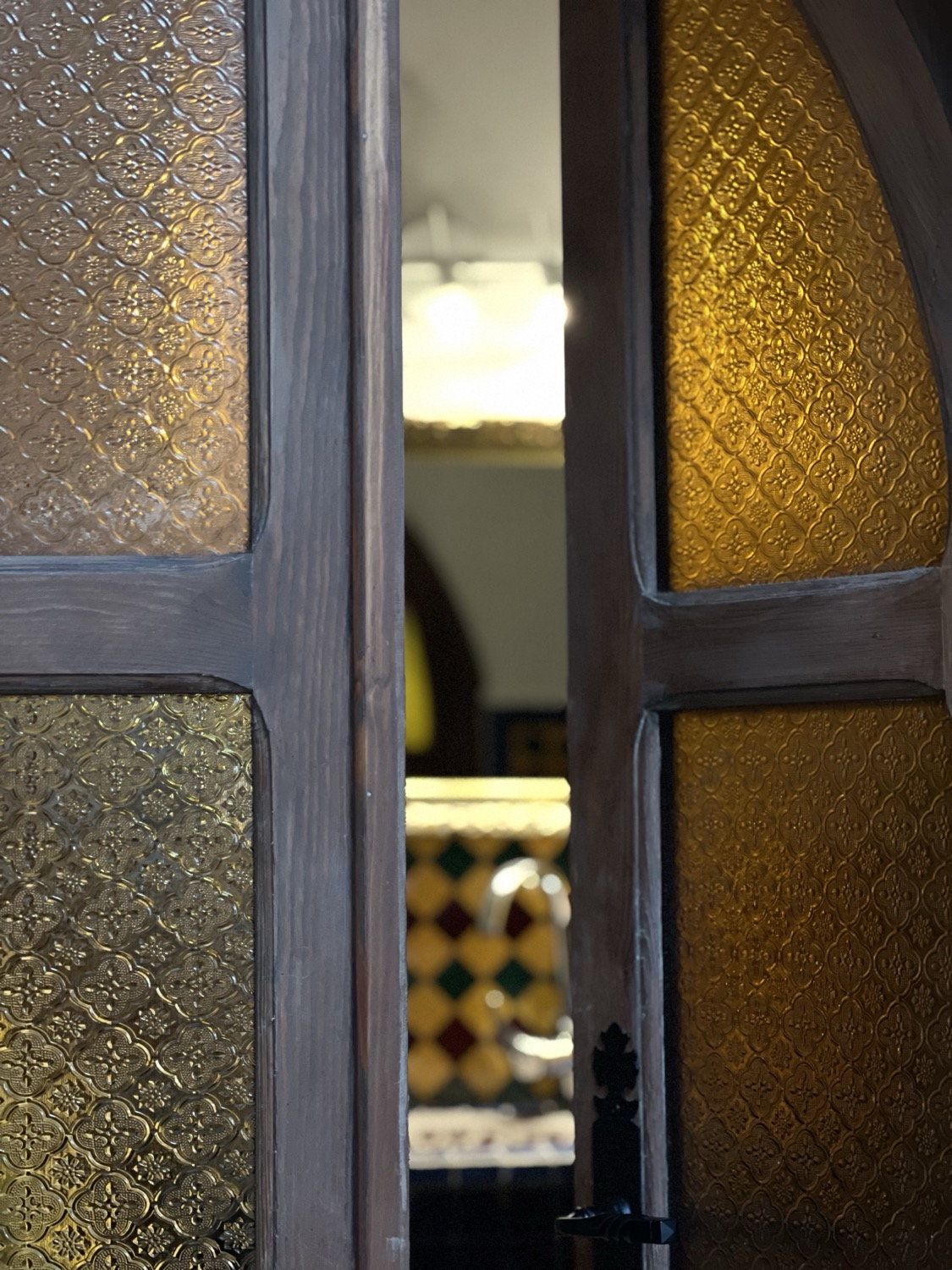 Ornate amber patterned glass door panels ajar, tiled floor and chandelier glimpsed beyond