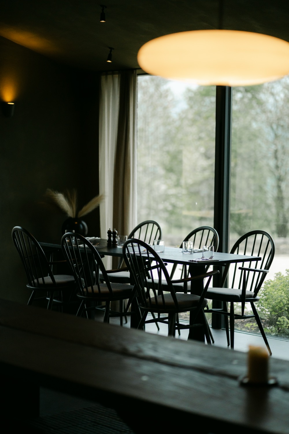 Dark restaurant interior with black Windsor chairs around tables, warm pendant light and large window onto woodland