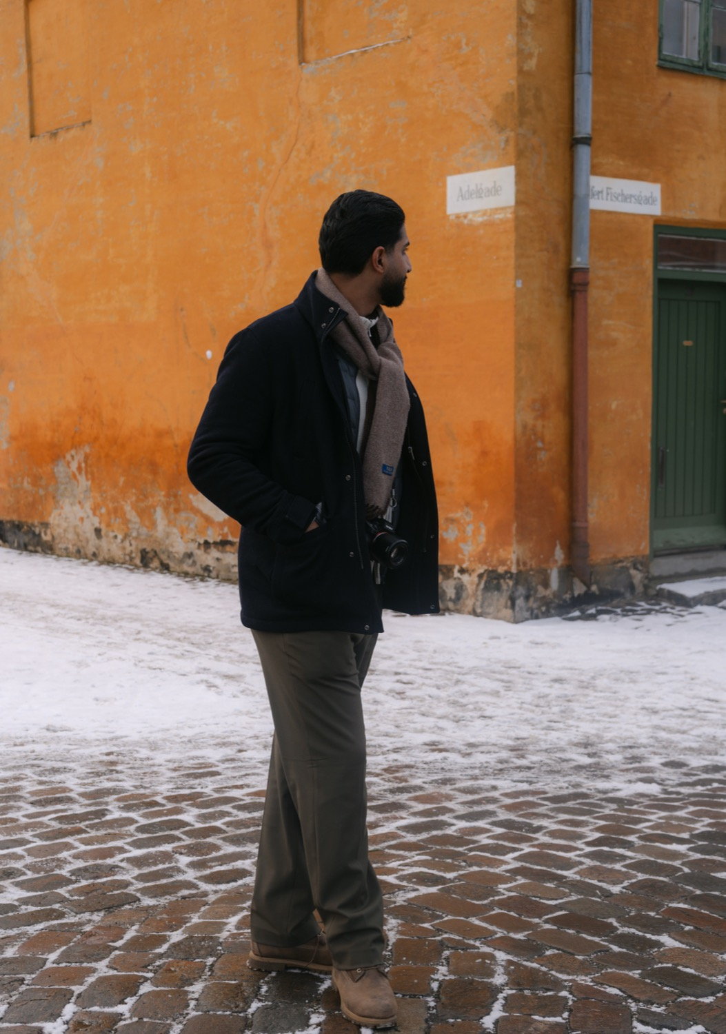 Man in dark jacket and scarf seen from the side against an orange-painted building, snow on cobblestones, Malmö