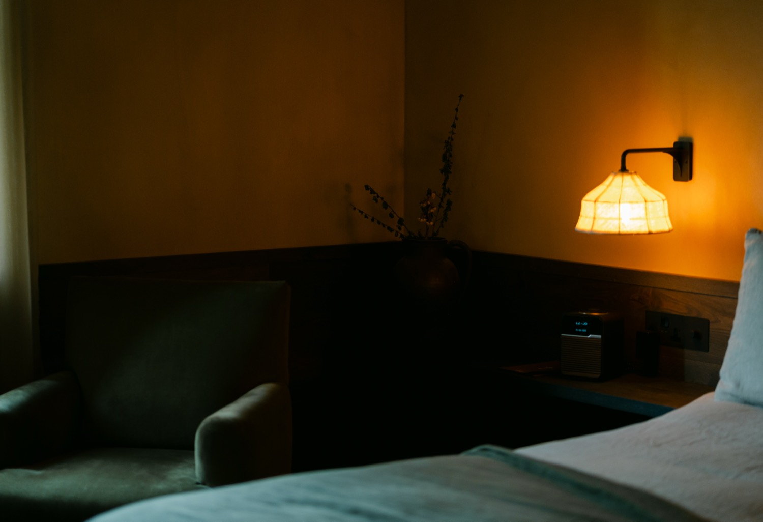 Dark hotel room corner at night, amber wall sconce glowing, velvet armchair and dried stems beside a clock radio
