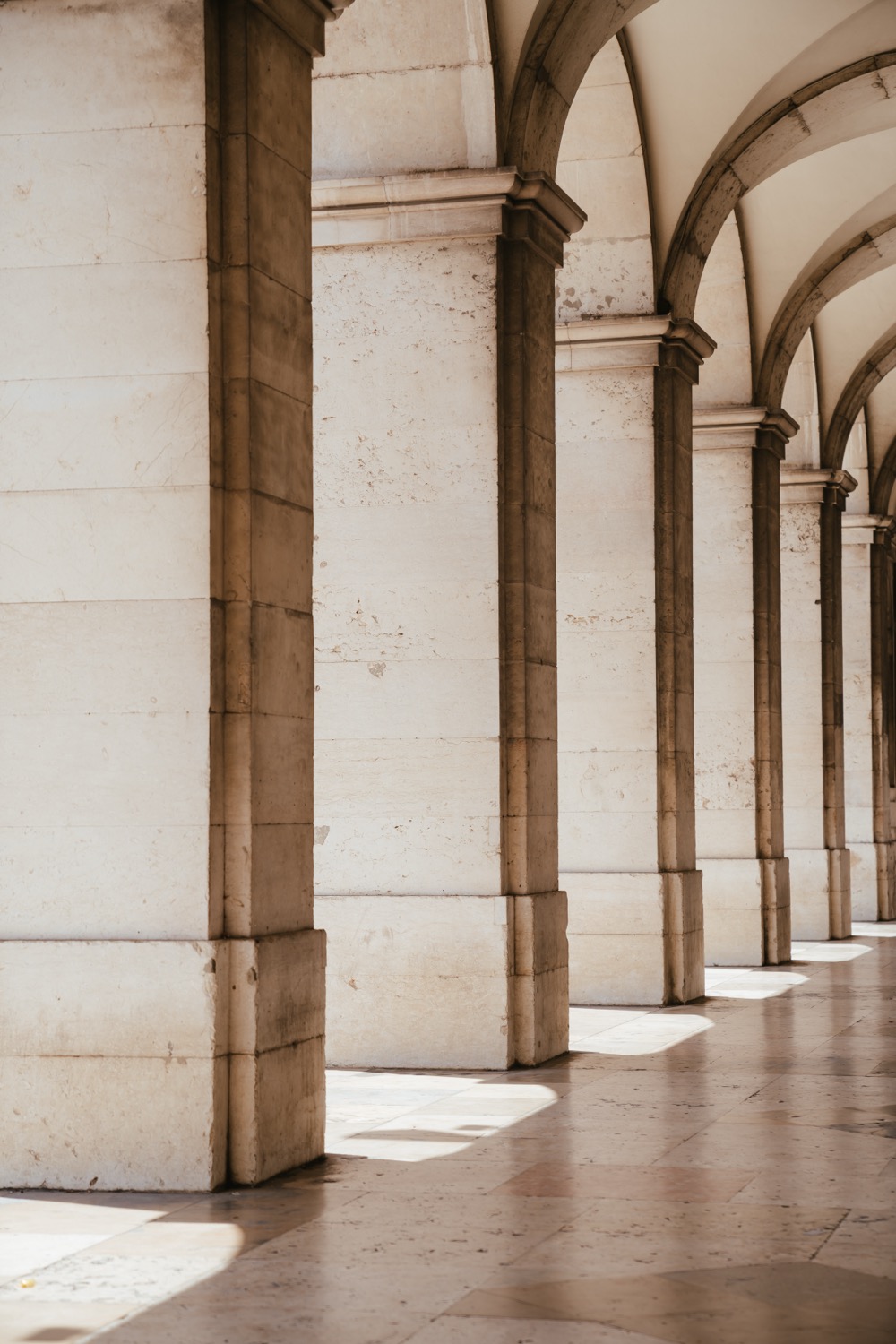 White stone colonnade with a row of arches receding in perspective, raking light on marble floor, Lisbon