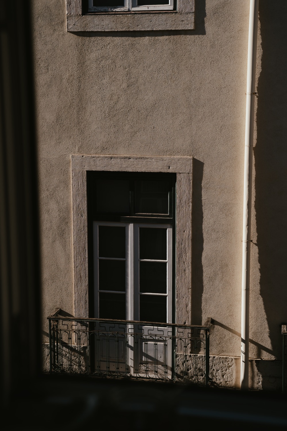 Plaster facade wall with deep raking shadow across it, tall window with iron balcony railing
