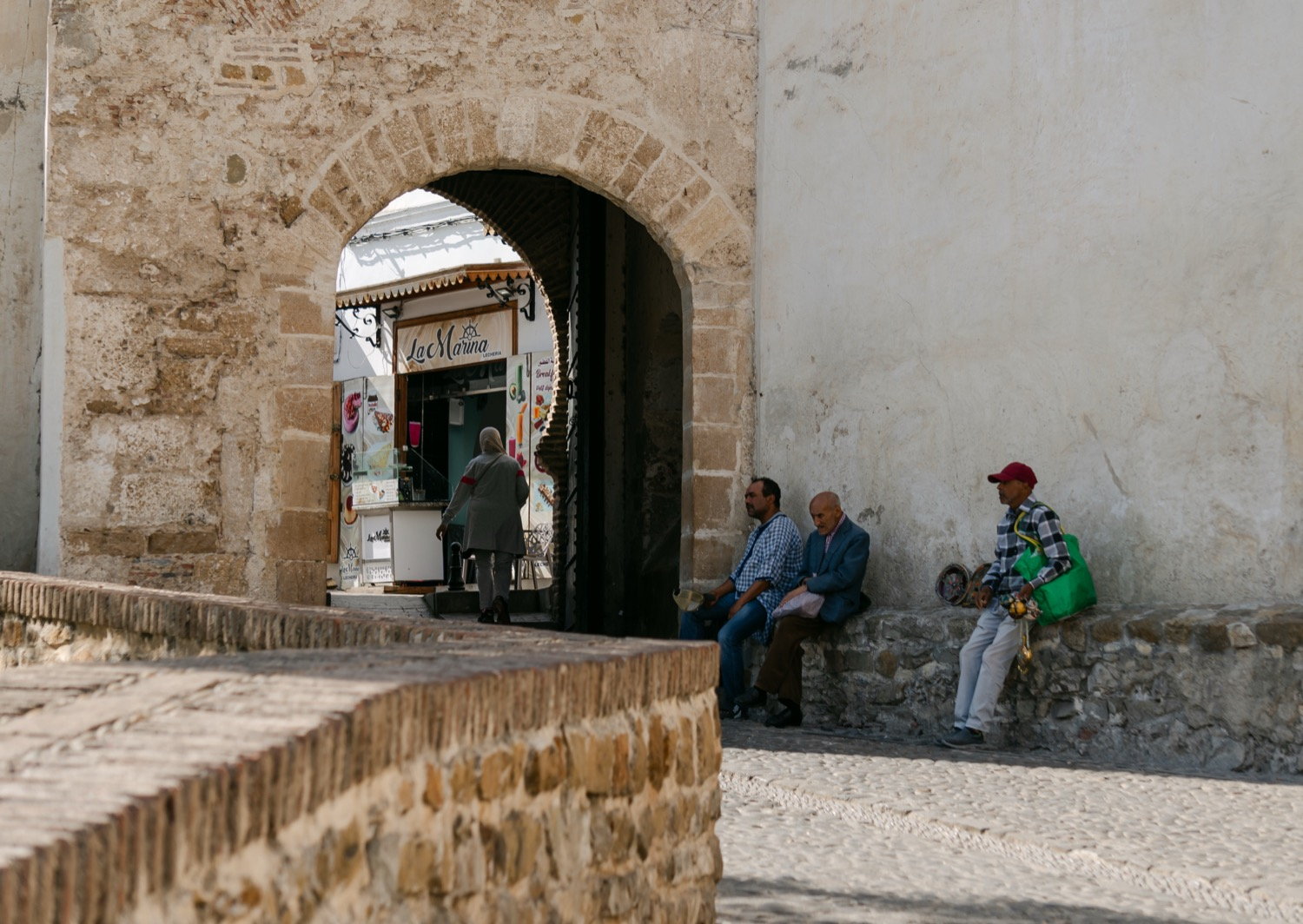 Three men resting against a stone wall under a medina arch, La Marina kiosk visible beyond, Tangier