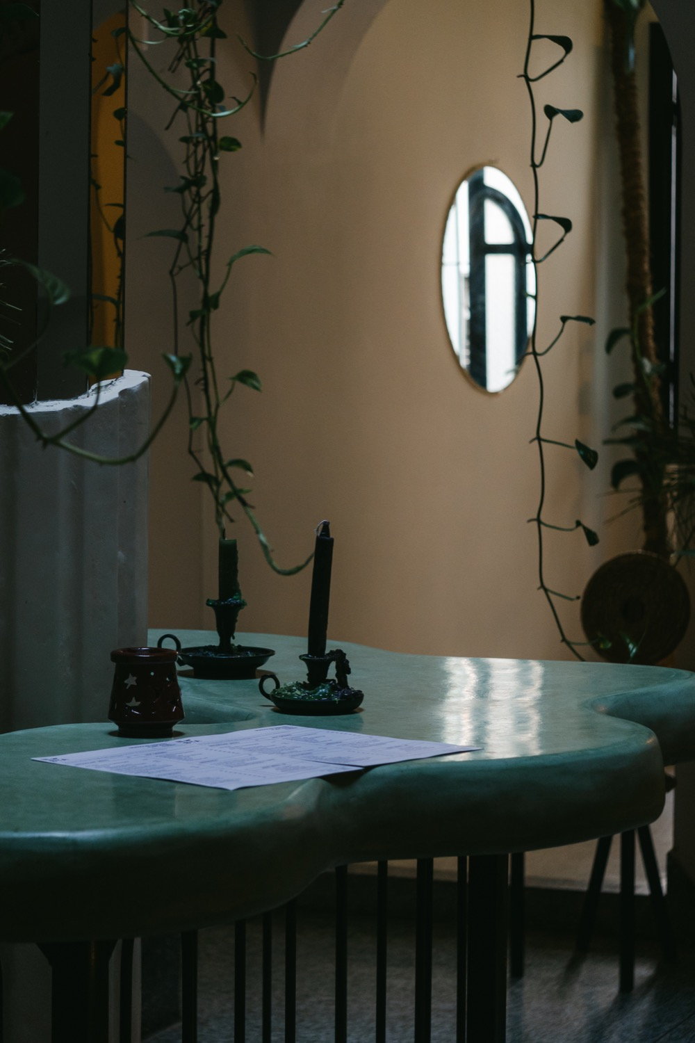 Green terrazzo table with two taper candles, trailing vines and a round mirror on a warm wall behind