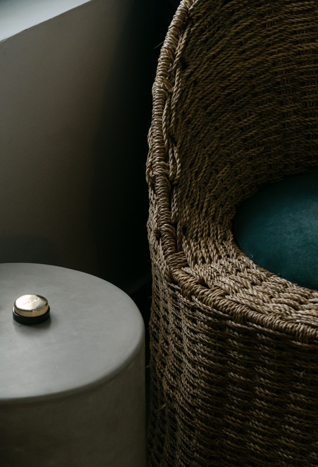 Close detail of a woven seagrass chair back with teal velvet cushion, dark concrete side table with brass-lidded candle beside it