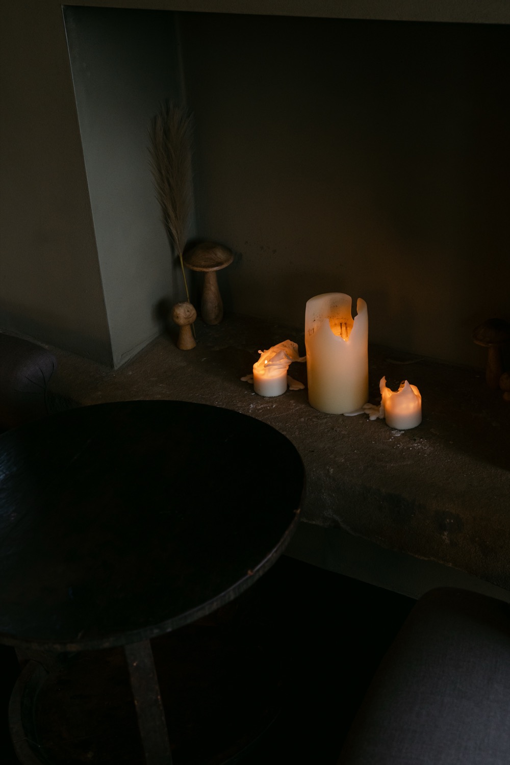 Pillar candle and votives on a low stone ledge beside a dark bathtub, pampas grass and carved wooden objects alongside