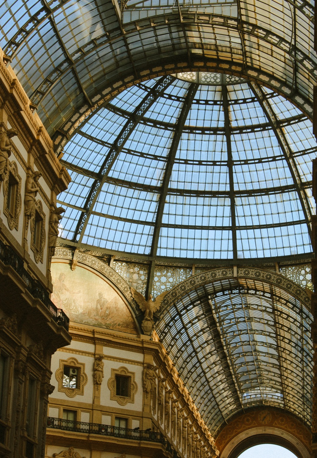 Looking up into the glass and iron dome of the Galleria Vittorio Emanuele II, gilded arched facades on either side, Milan