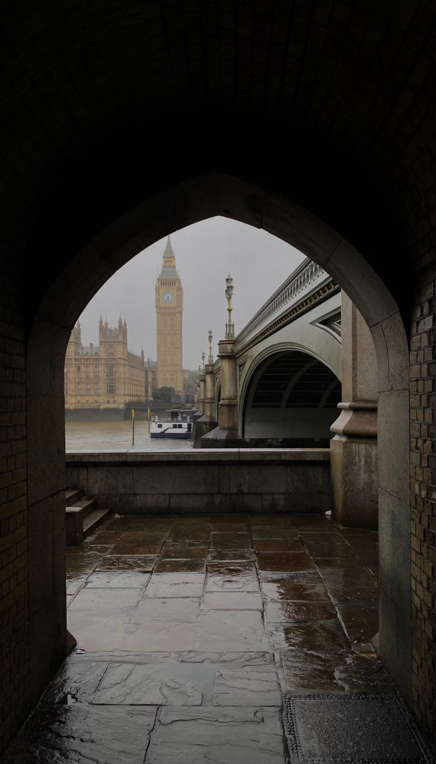 Big Ben and the Houses of Parliament seen through a Gothic stone arch at Westminster, rain-wet paving stones in foreground, London