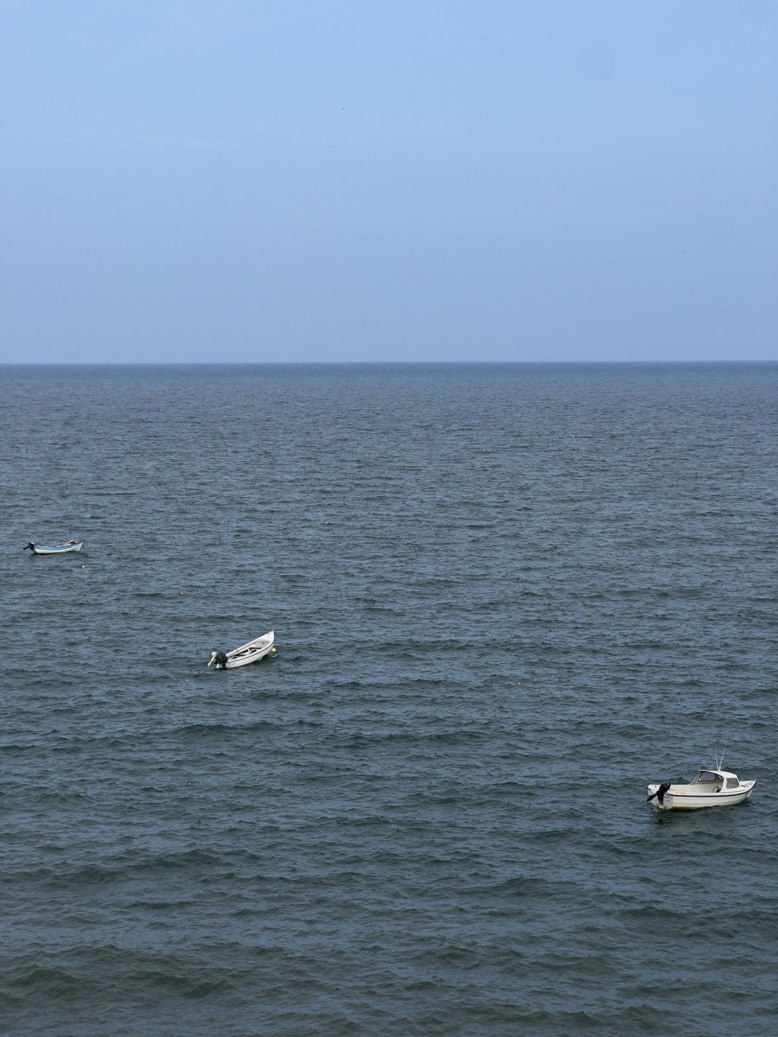 Three small white boats moored on calm grey-blue open water viewed from above, pale sky at horizon