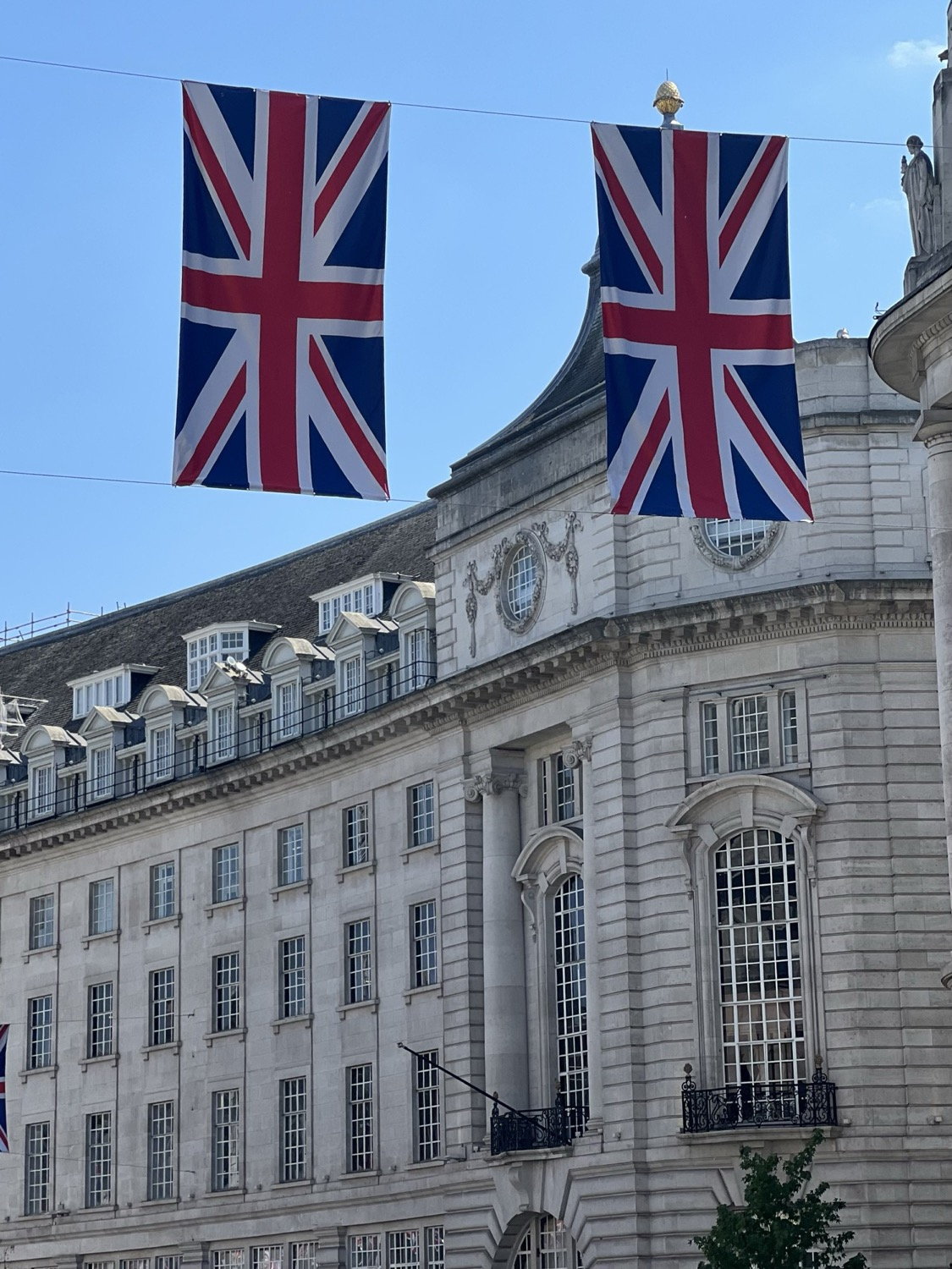 Two Union Jack flags hanging over Regent Street, neoclassical curved facade behind, blue sky, London