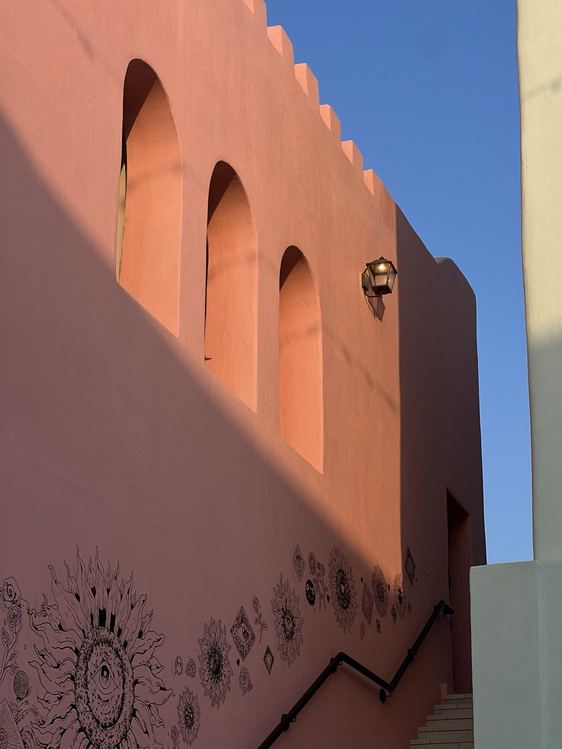 Salmon-pink building facade with arched recesses, battlemented parapet and celestial sun illustration painted on lower wall, golden light, Marrakesh