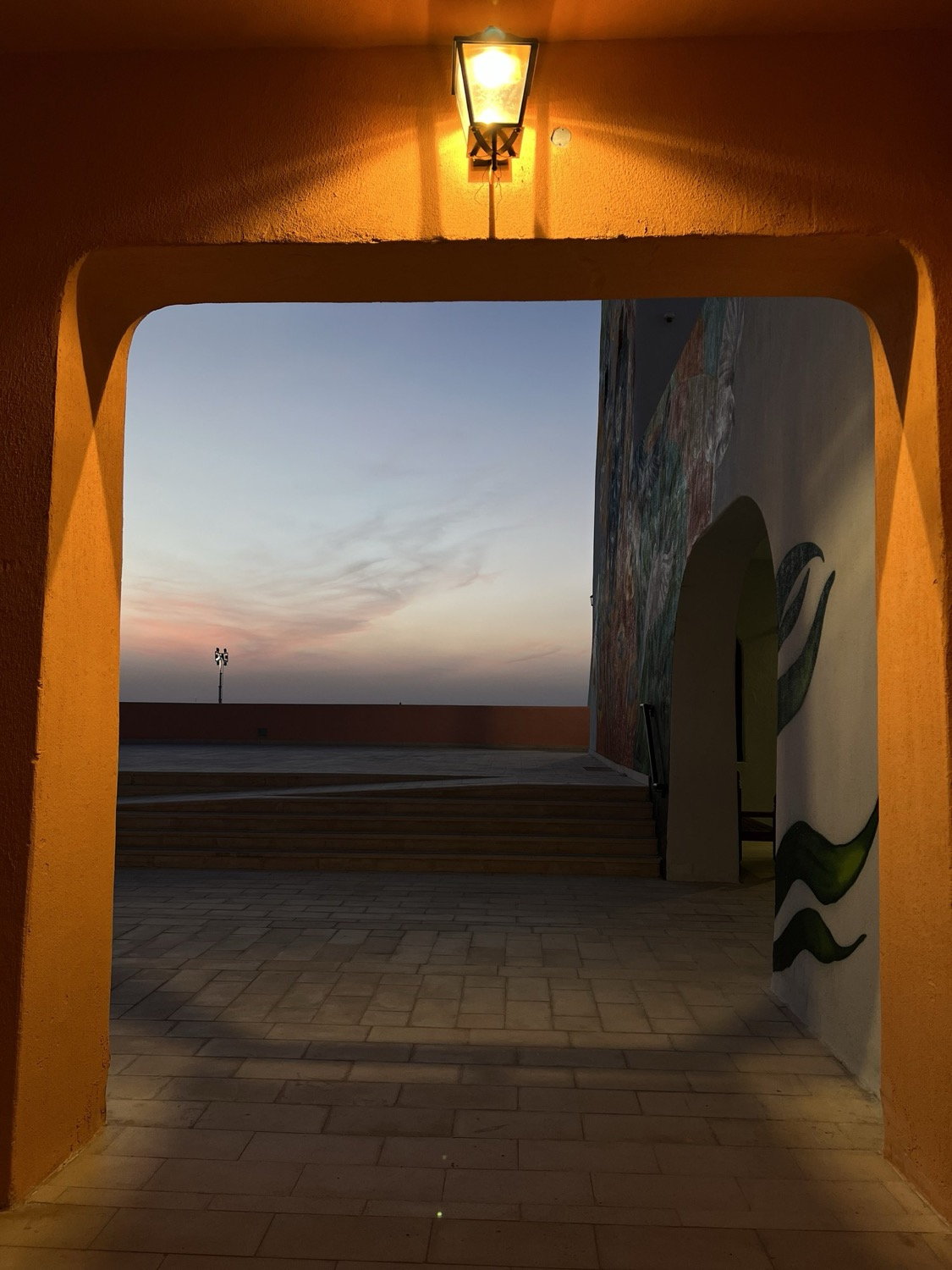 Orange-plastered archway with lantern above, looking through to a pastel dusk sky and painted mural on the right wall, Asilah