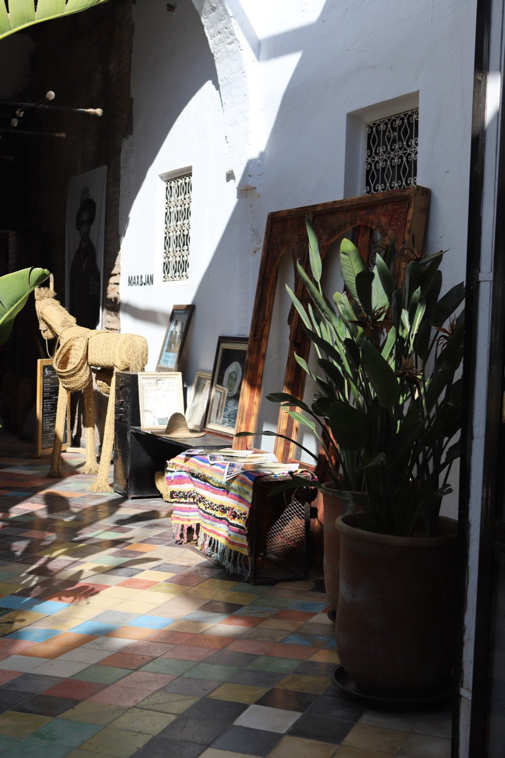 Gallery courtyard with colourful zellige tile floor, straw horse sculpture, tropical plant and framed works, dappled sunlight, Tangier