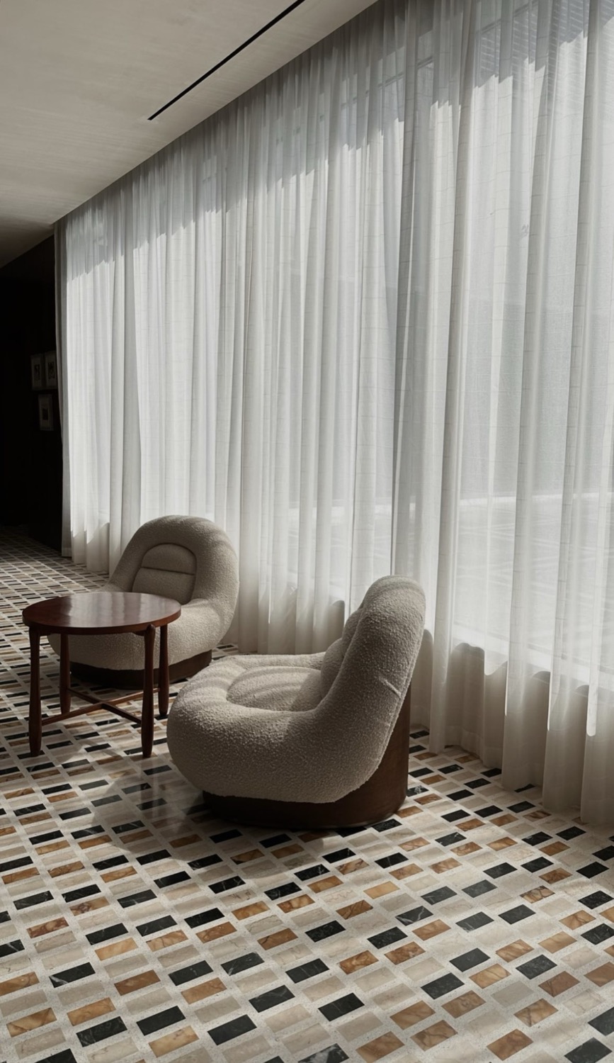 Two boucle lounge chairs beside a round table, sheer floor-to-ceiling curtains and terrazzo floor