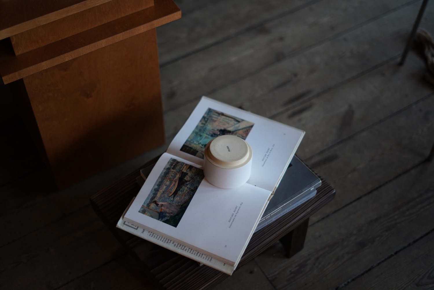 Art book open to Impressionist paintings, small white ceramic pot resting on the pages, warm oak floor