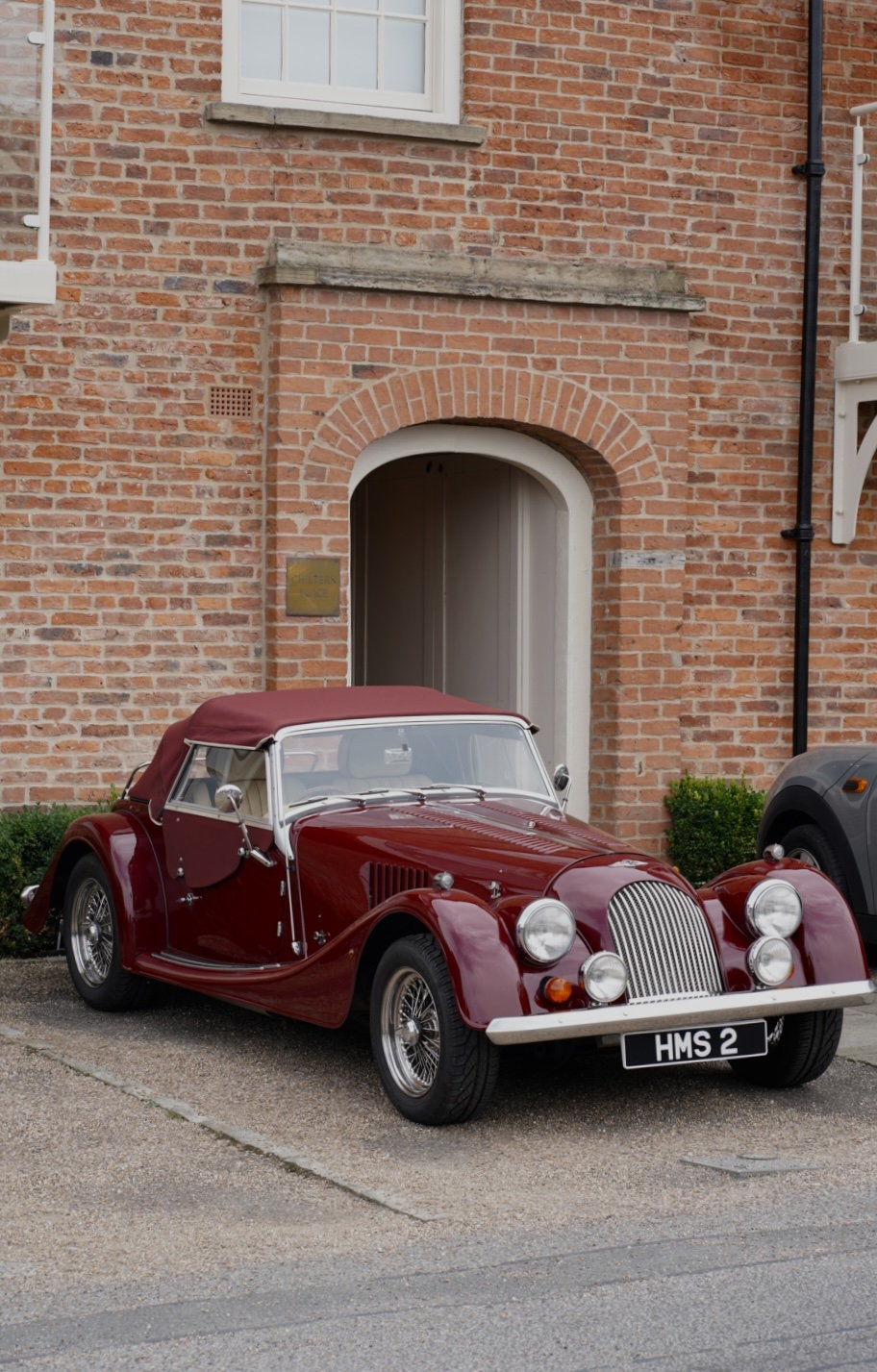 A burgundy Morgan roadster parked before a brick facade