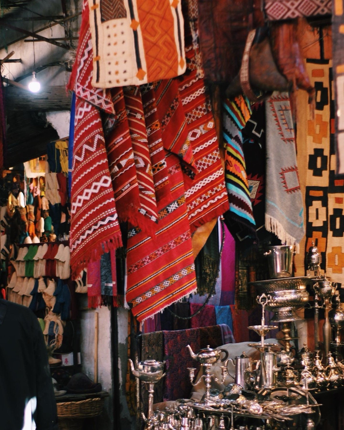 Red and orange Berber textiles hanging in a Marrakesh souk, silver teapots below