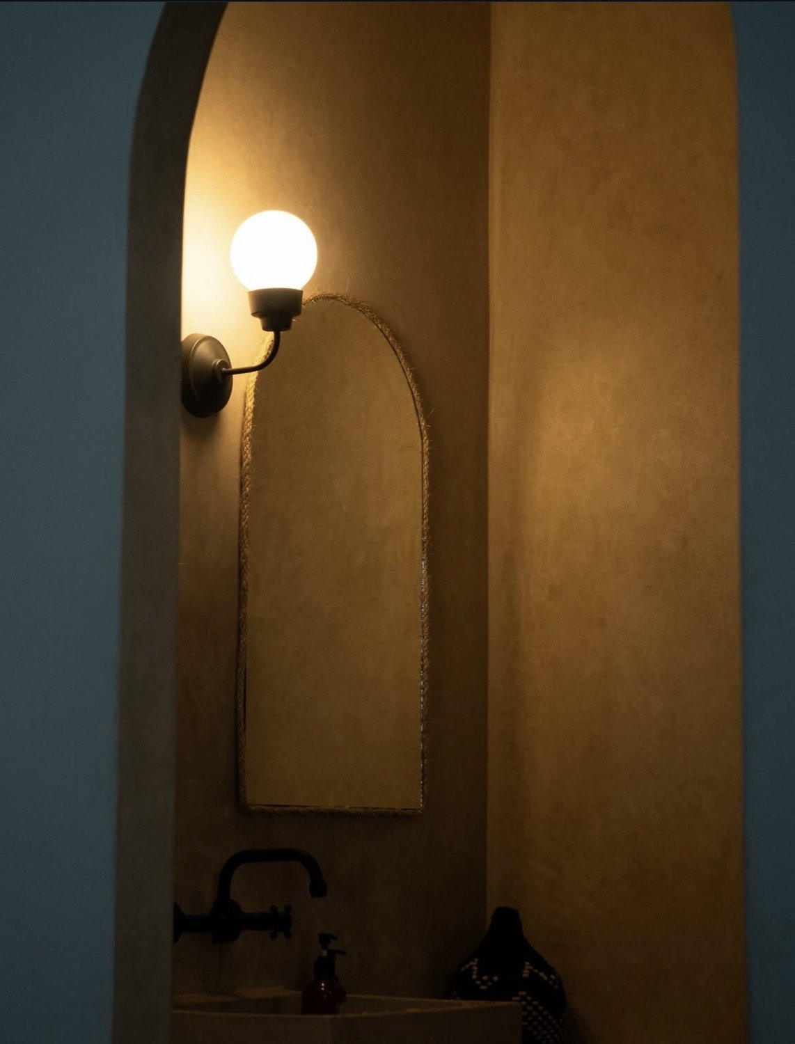 Warm plaster arched alcove with globe wall sconce, arched mirror and black tap below