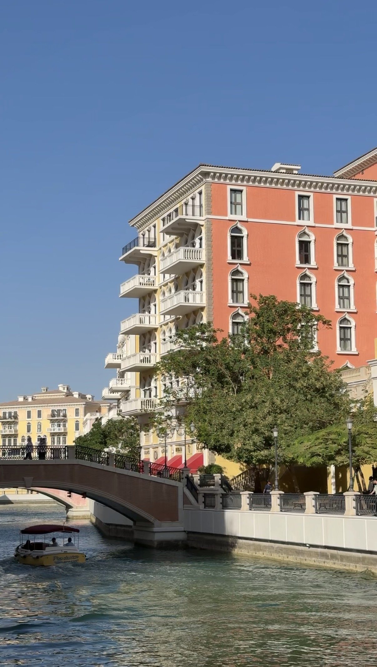 A coral apartment building beside a bridge and passing boat, Qanat Quartier, Doha