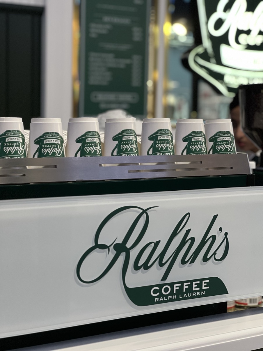 Row of branded Ralph's Coffee cups lined up on a white counter with the script logo below