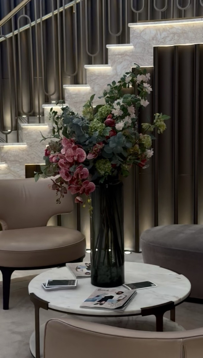 Large floral arrangement in a dark glass vase beside soft seating and a lit staircase, Salon de Parfums, Harrods, London