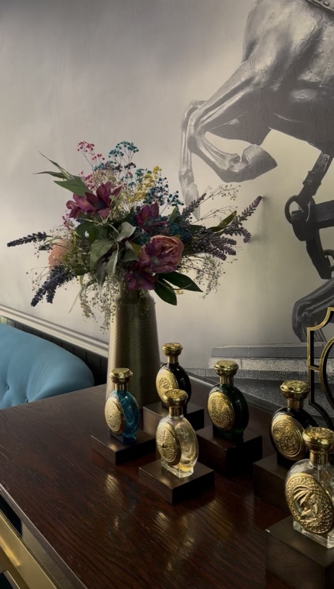 Ornate gold-capped perfume bottles beside a tall flower arrangement on a polished dark table, Salon de Parfums, Harrods, London